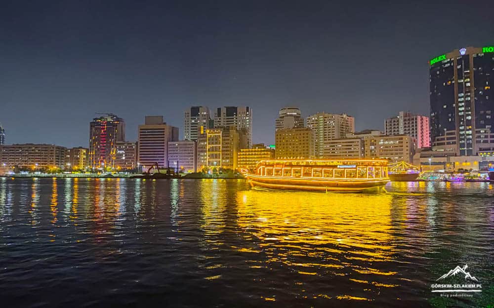 Nighttime city skyline along a river with a bright yellow-lit boat and reflections on the water.