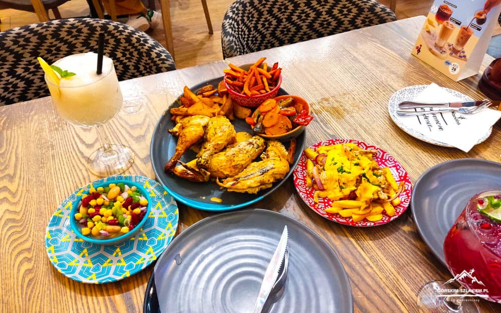 Shared meal on a wooden table: roasted chicken, potato wedges, cheesy fries, and a colorful bean salad with a yellow cocktail on the side.