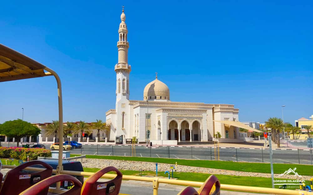 White mosque with dome and tall minaret under a clear blue sky, palm trees and outdoor seating in foreground.