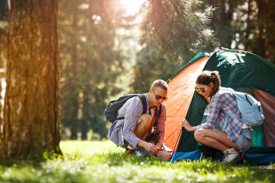Two hikers setting up a green and orange tent in a sunlit forest clearing.