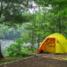 Bright yellow tent pitched on a lakeside shore among tall green trees and calm water nearby.