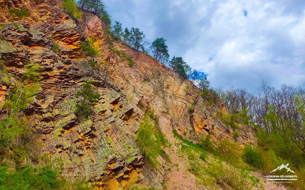 Steep sedimentary rock cliff with orange-gray layers, sparse trees, and a cloudy blue sky above.