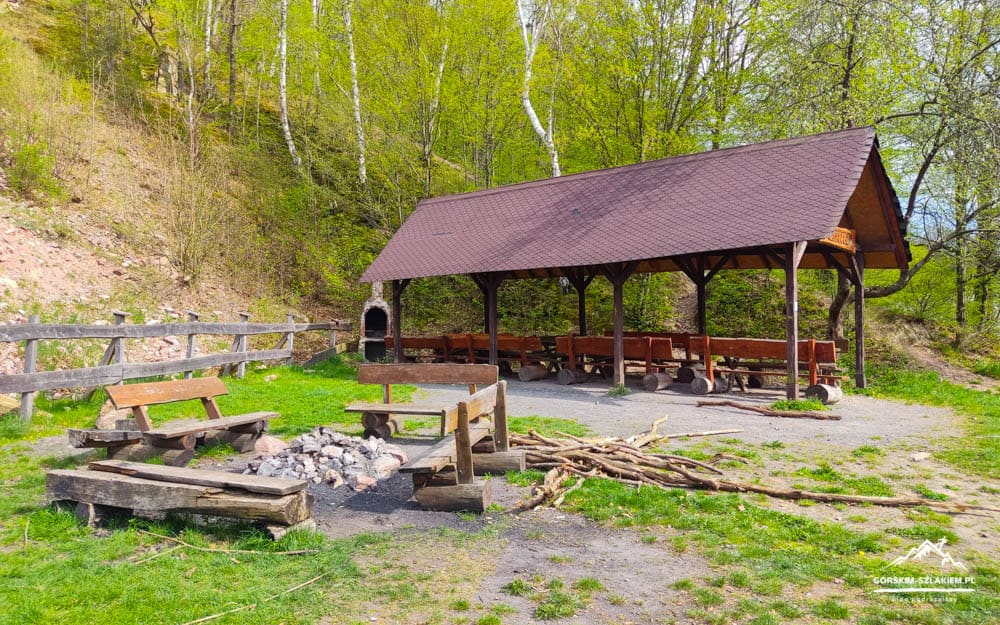 Wooden pavilion with long bench seating beside a campfire pit in a forested clearing.