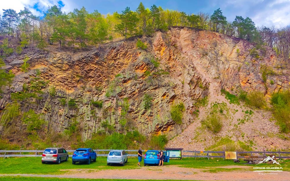 Steep rocky cliff with trees at the top; cars parked along a fence and a few people near a blue car by the lot.