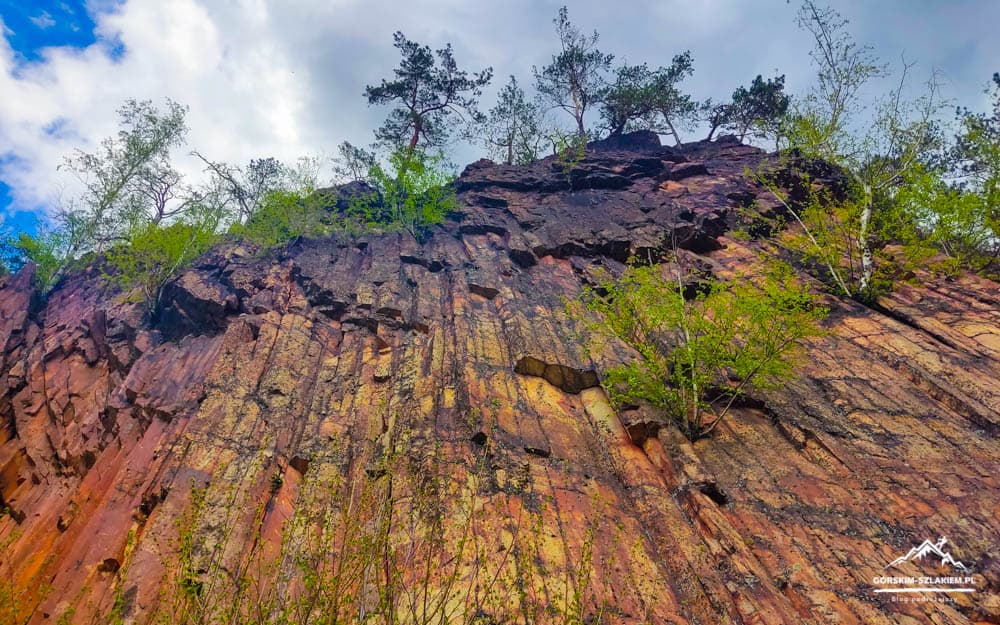 Colorful, stratified cliff face with small trees at the top and a blue sky above.