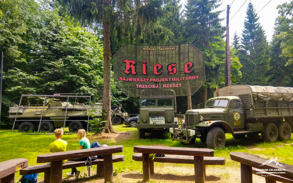 Outdoor park scene with vintage military trucks and a large wooden sign reading 'Riese' in red letters, set among trees; two children sit on a bench in the foreground.