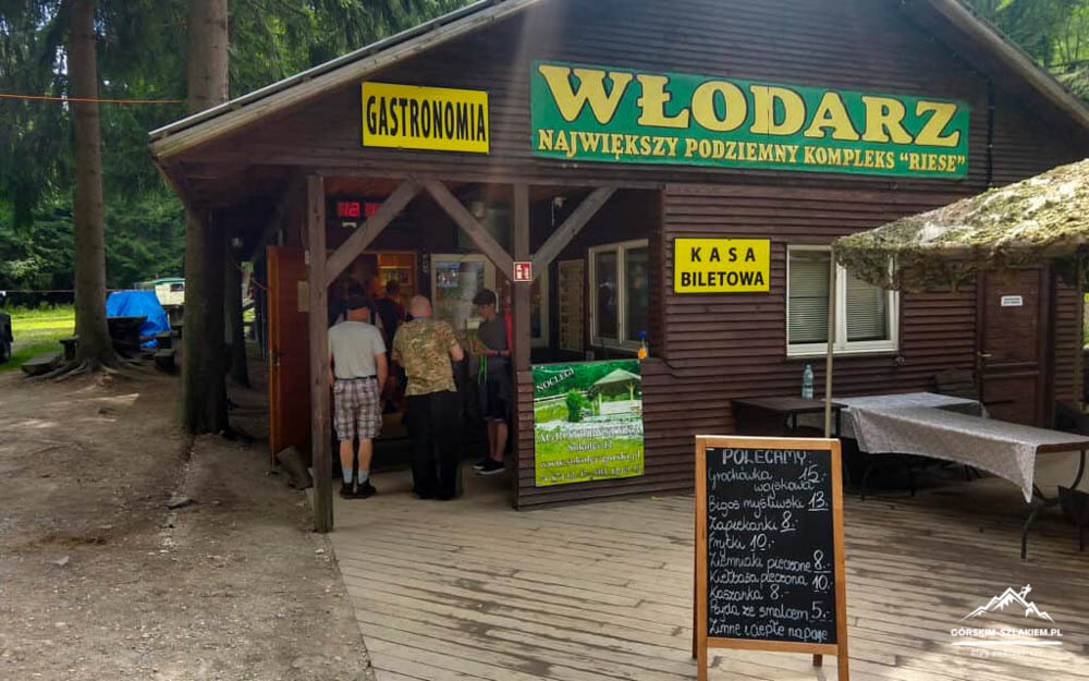 Wooden rustic kiosk in a forest with a line of people at the counter; signs read 'Gastronomia' and 'Włodarz' (big green-yellow sign) and 'Kasa Biletowa' nearby, chalkboard menu outside.
