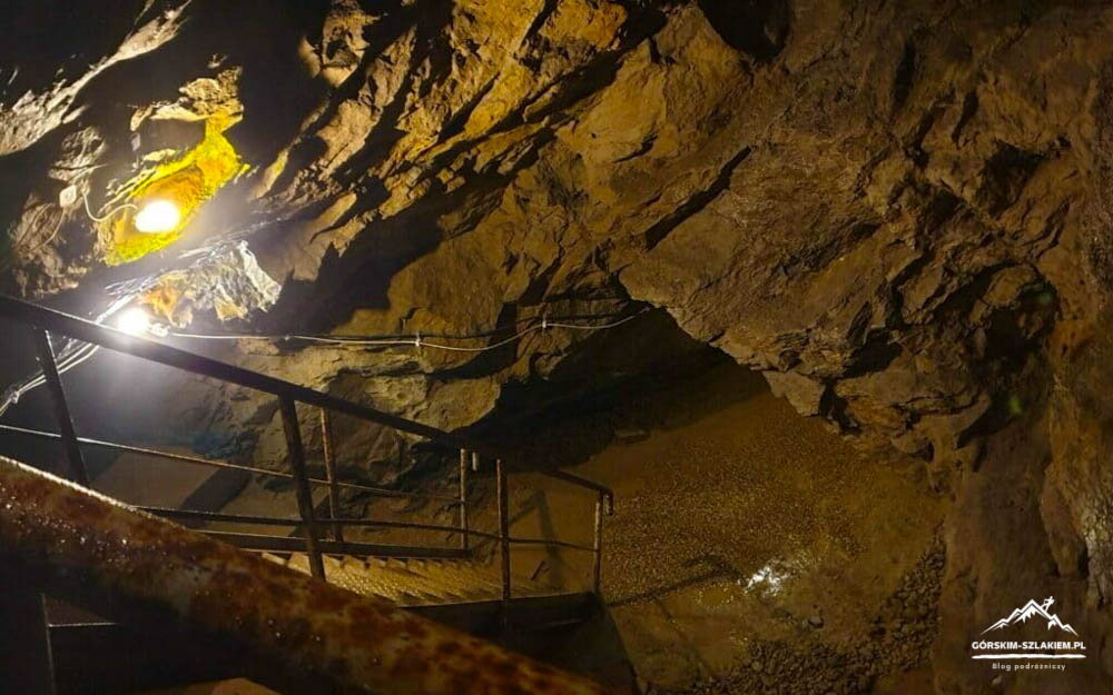 Interior of a dim cave with a metal staircase and railing, illuminated by overhead lights; wet rocky walls and a small pool at the bottom, with a logo watermark in the lower-right corner.