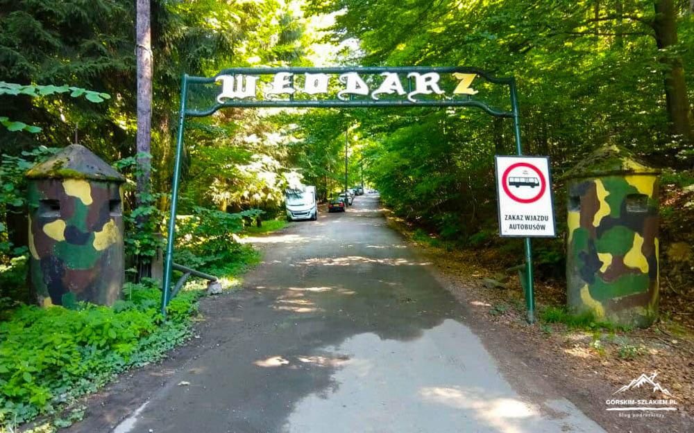 Forest road entrance framed by camouflaged pillars and a green metal arch gate in a forest setting, with a 'no buses' sign nearby