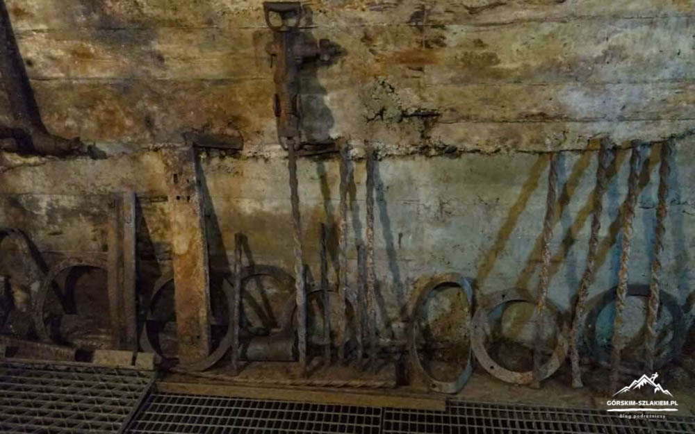 Damp, rusty mine interior with hanging ropes and circular iron rings along the wall, and a grated floor in the foreground.