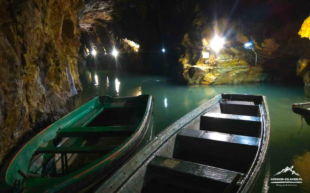 Two wooden boats rest on a turquoise underground lake inside a rocky cavern, illuminated by artificial lights.