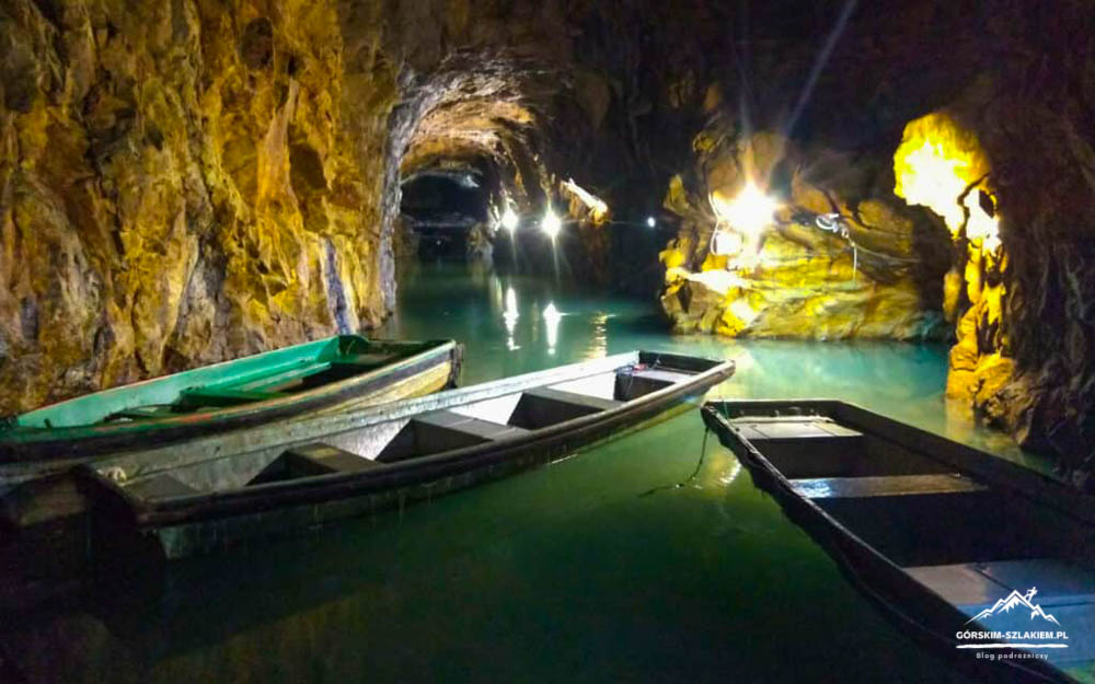 Inside a dim cave, turquoise water with several wooden boats moored along rocky walls, illuminated by artificial lights.