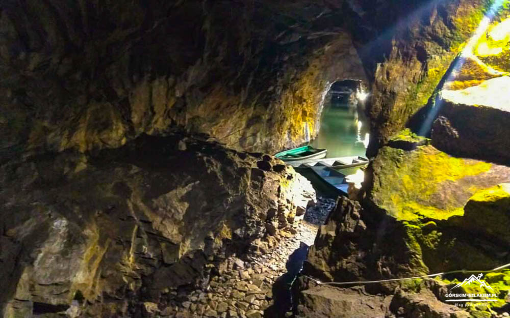 Inside a rocky cave with a narrow water-filled tunnel and small boats docked along the shore.