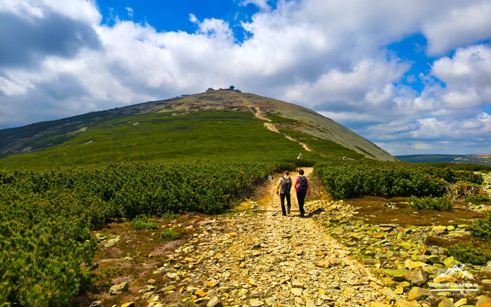 Two hikers with backpacks walk along a rocky trail toward a grassy hill under a bright, cloudy sky.