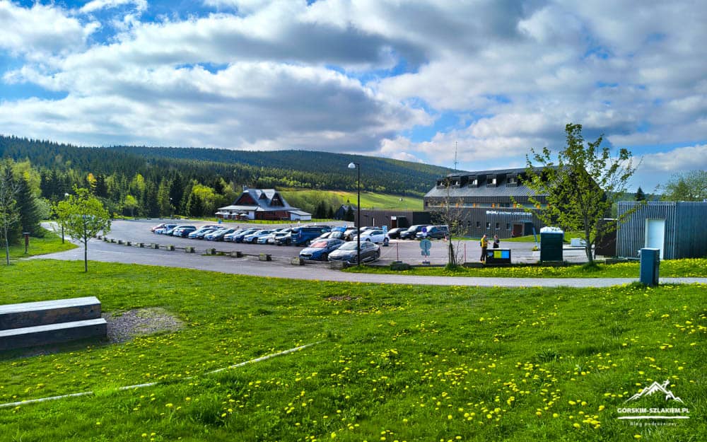 Parking lot with many cars next to a lodge and a modern building, set in a green hillside landscape under a cloudy blue sky.