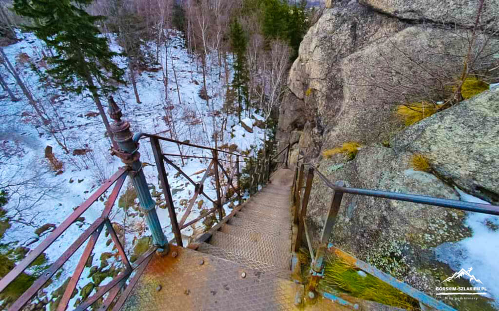 Rusty metal staircase clinging to a rocky cliff, railing on both sides, descending toward a snow-covered forest below.