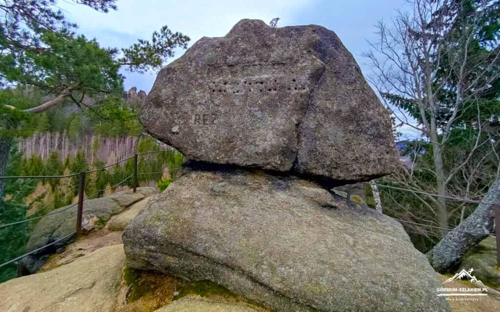 Stacked boulder balanced on a larger rock along a trail with a railing and forest in the background.