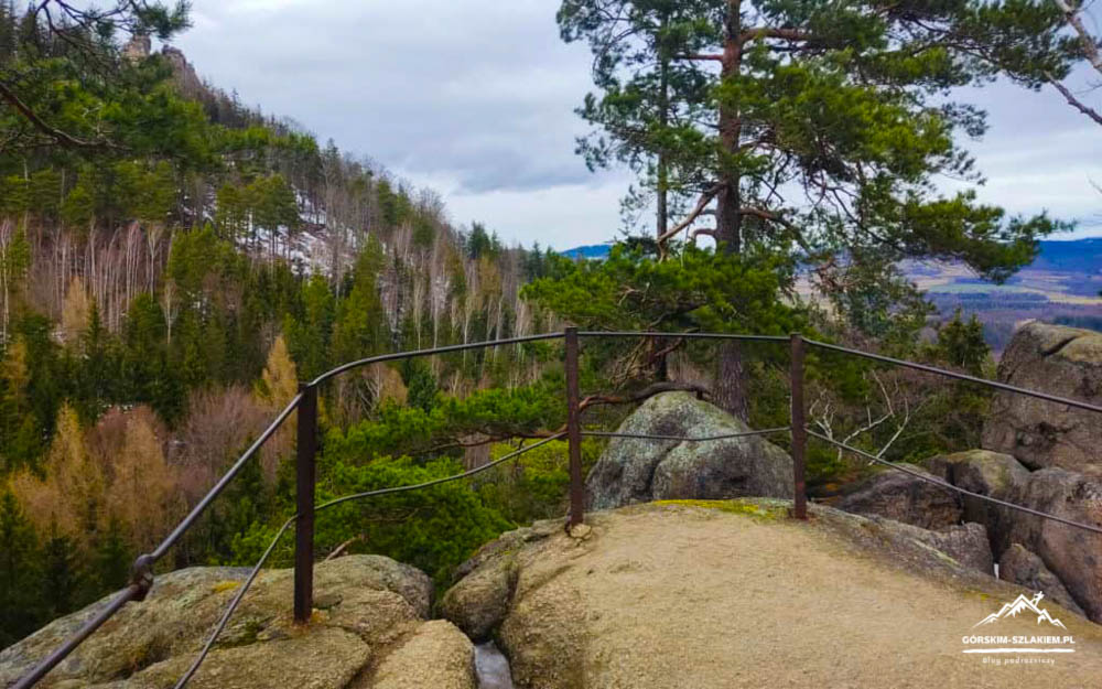 Rocky cliff edge with a curved metal railing, overlooking a pine forest valley and distant hills on a cloudy day.