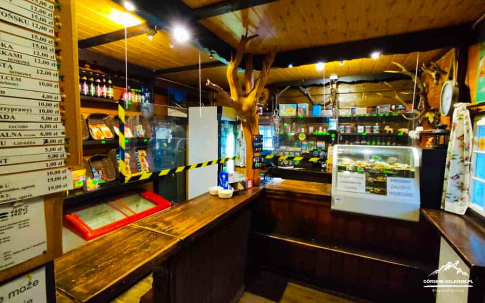 Cozy rustic shop interior with a wooden counter, glass display case of deli items, shelves of products, and a tree centerpiece; yellow/black caution tape blocks off a section.