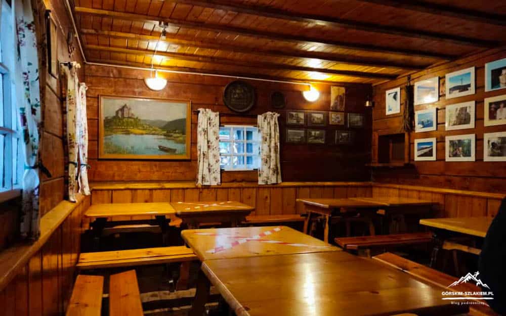 Cozy rustic dining room in a wood-paneled cabin with wooden tables and benches, framed painting, and wall photos under warm lights, curtains at windows.