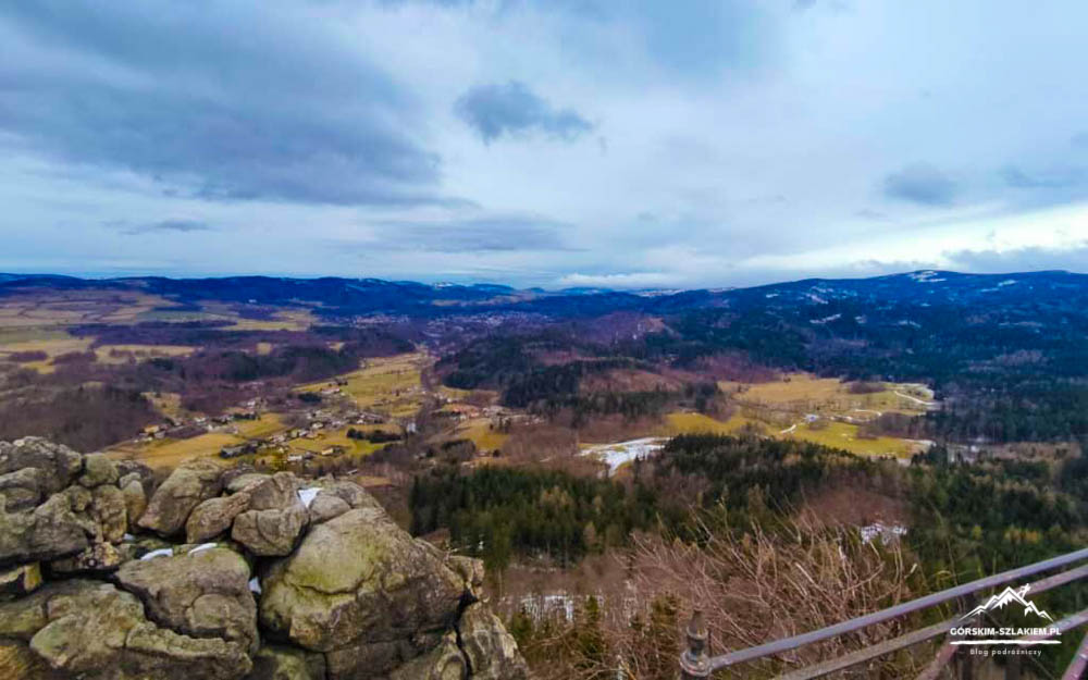 Rocky stone foreground with a metal railing frames a wide valley of fields and forests under a cloudy sky, distant hills on the horizon.