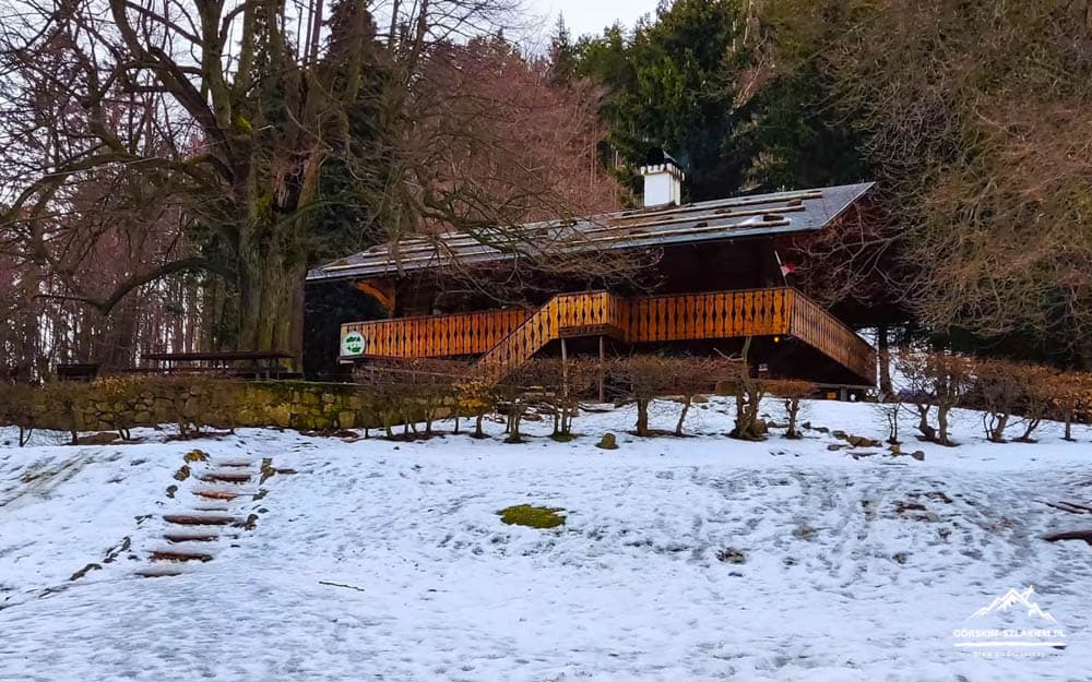 Snowy hillside with a wooden chalet on stilts and a wraparound balcony, surrounded by bare trees.