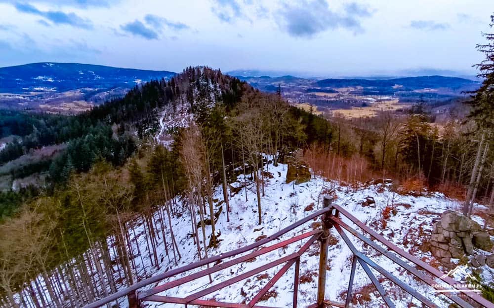 Snowy hillside viewpoint with a metal railing deck overlooking a forested valley and distant hills.