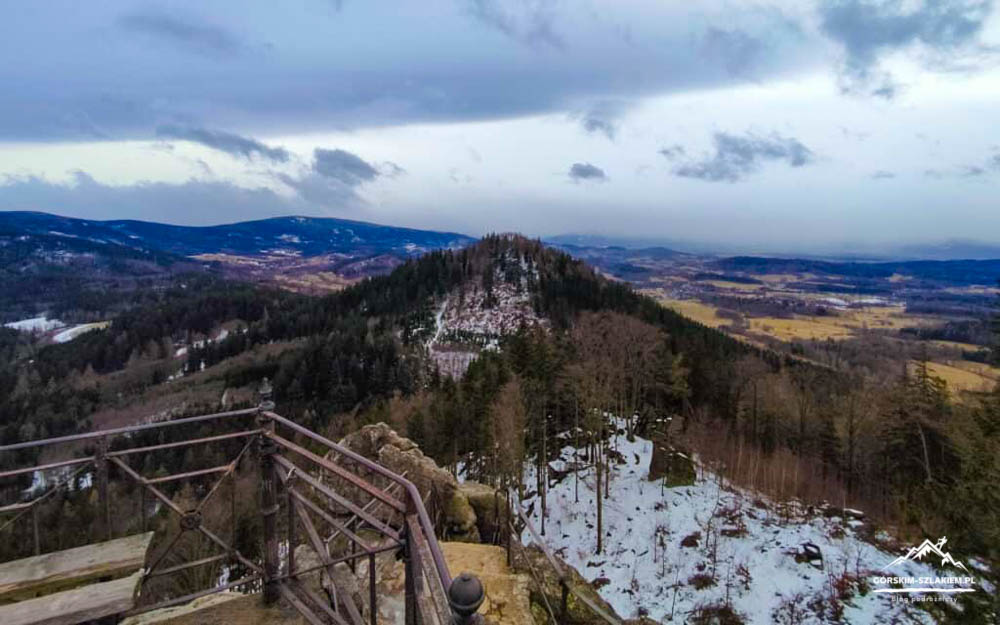 Snow-covered hillside viewpoint with a metal railing in the foreground, evergreen trees, and a distant valley under a cloudy blue sky.