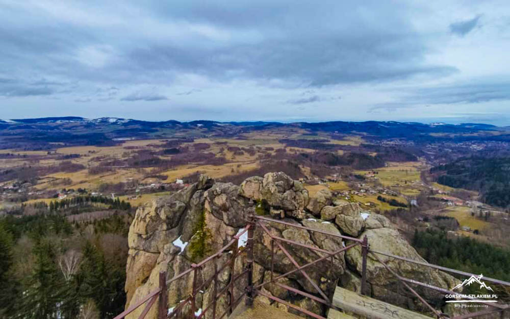 Rocky viewpoint with a metal railing, overlooking a valley and hills under a cloudy sky.