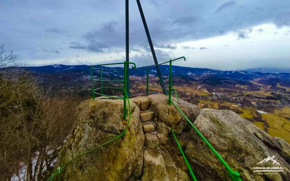 Rocky cliff overlook with bright green railing and stone steps, gazing over a valley and distant hills on a cloudy day.