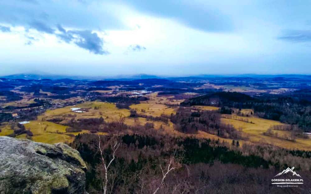 Panoramic valley view from a rocky overlook: patchwork fields, forests, and distant hills under a cloudy blue sky.