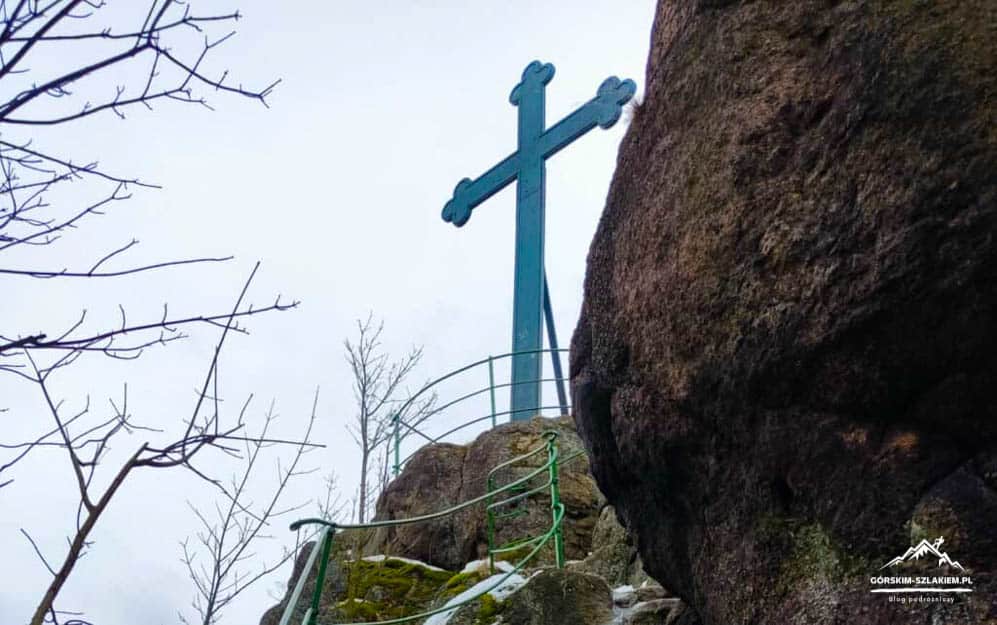 Large blue metal cross mounted on a rocky cliff edge, with a winding green railing and patches of snow on the ground.","Blue cross sculpture atop a rocky overlook, near bare trees and a metal railing along a narrow path.","Religious cross on a mountain rock face overlooking the landscape in a wintry setting.","Cross on the edge of a cliff with safety railing along a rocky trail.","Blue cruciform structure at a high overlook with bare branches and rugged rock behind it."