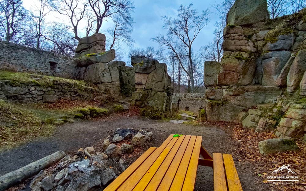 Stone ruins with large boulders and a bright wooden picnic table in a leafless forest setting.