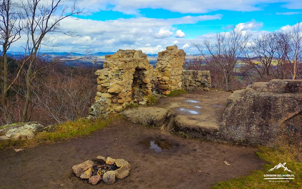 Ruined stone archways on a hilltop overlook with a dirt path, puddles, and a distant valley under a bright blue sky.