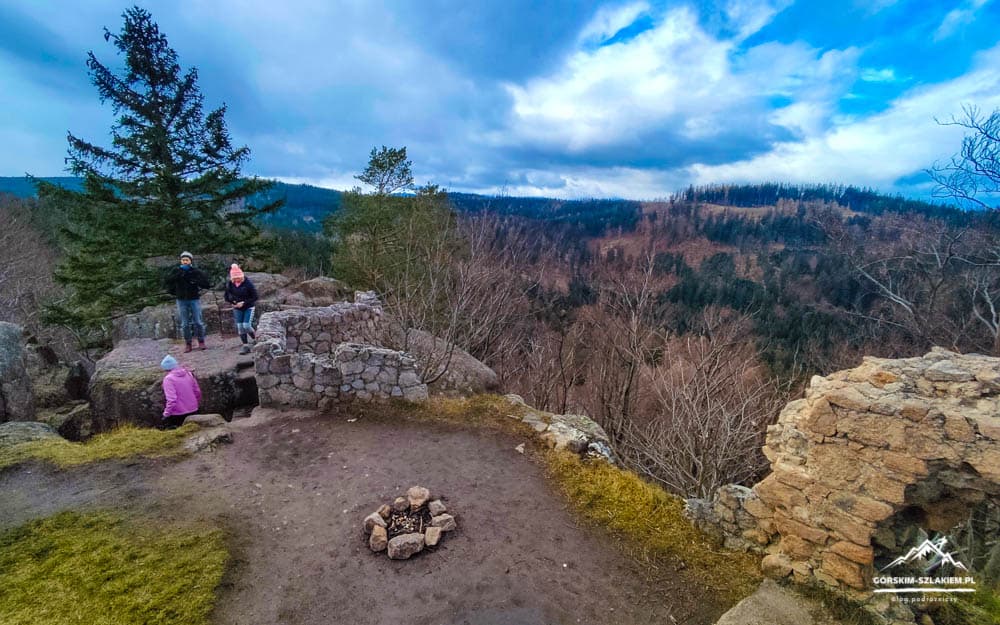 Three hikers stand on a rocky overlook with a stone wall and fire pit, gazing over a forested valley beneath a cloudy sky.