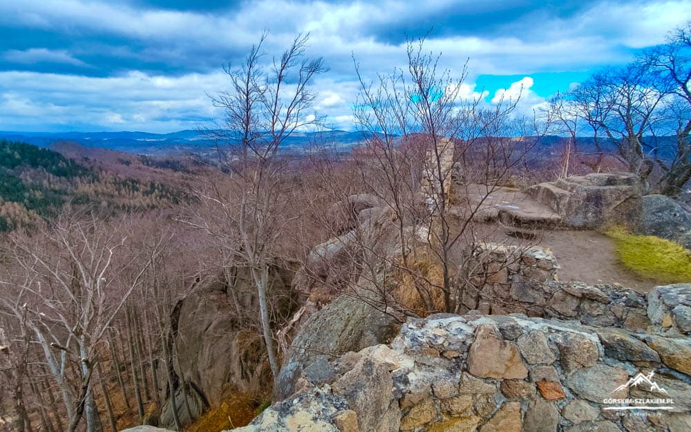Rocky cliff overlook with a stone wall, bare trees, and a distant forested valley under a cloudy blue sky in winter.