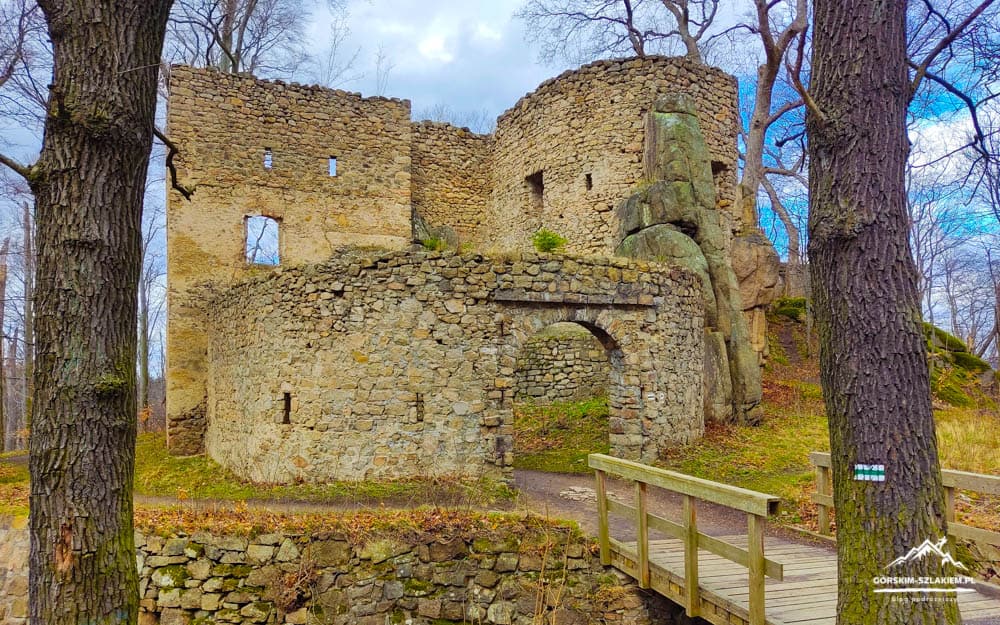Stone fortress ruins with an arched doorway, surrounded by leafless trees and a wooden footbridge