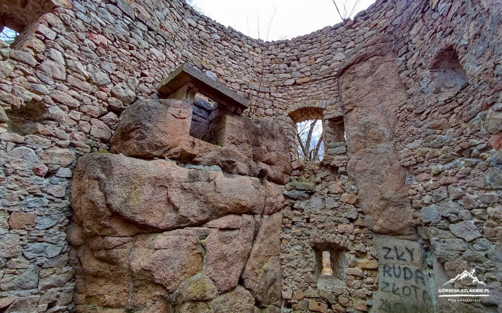 Interior of a circular stone ruin with large boulder walls, small arched openings, and graffiti on a pillar; leafless trees visible through a doorway in the distance.