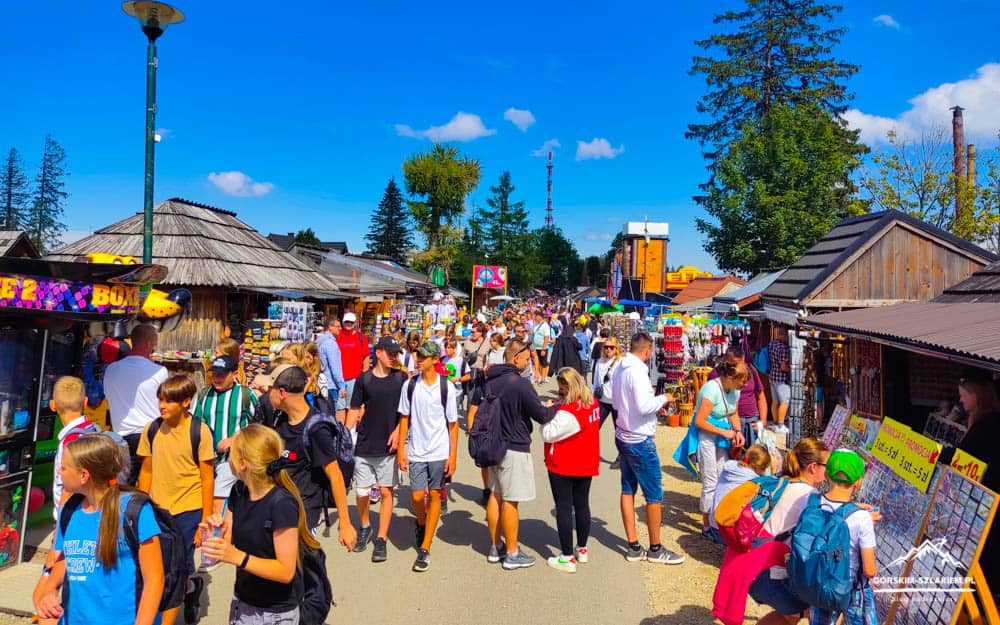 Crowded outdoor market with wooden stalls, colorful signs, and shoppers on a sunny day.