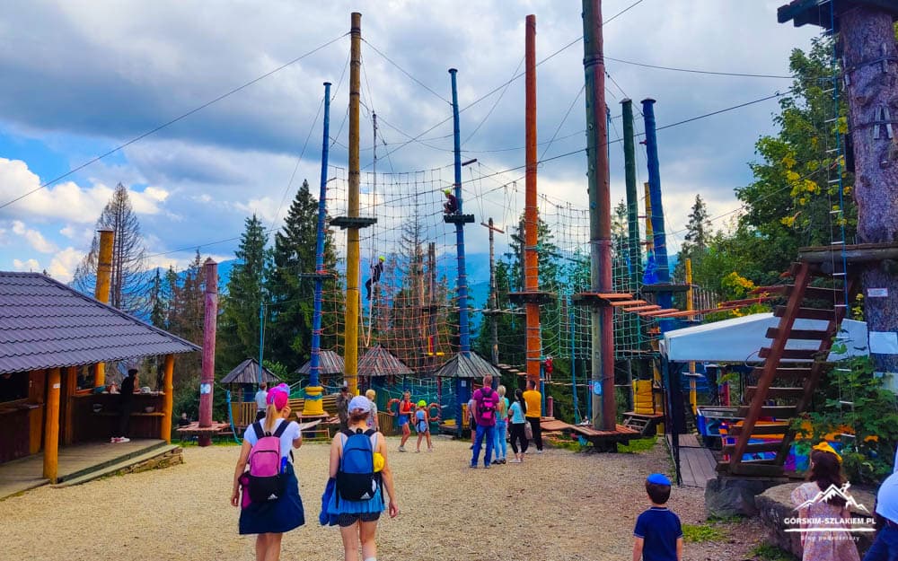 Children with backpacks walk toward a colorful multi‑tower ropes course with nets and wooden platforms in a forest park area.