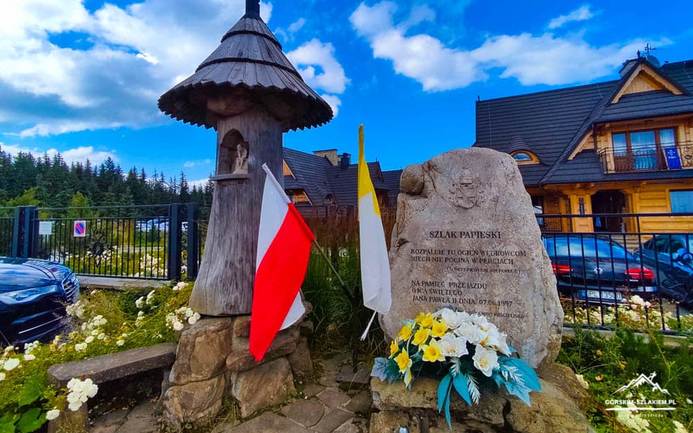 Memorial stone with inscription, two flags (Poland flag and white-yellow flag), and bouquet of flowers beside a wooden shrine under a blue sky.
