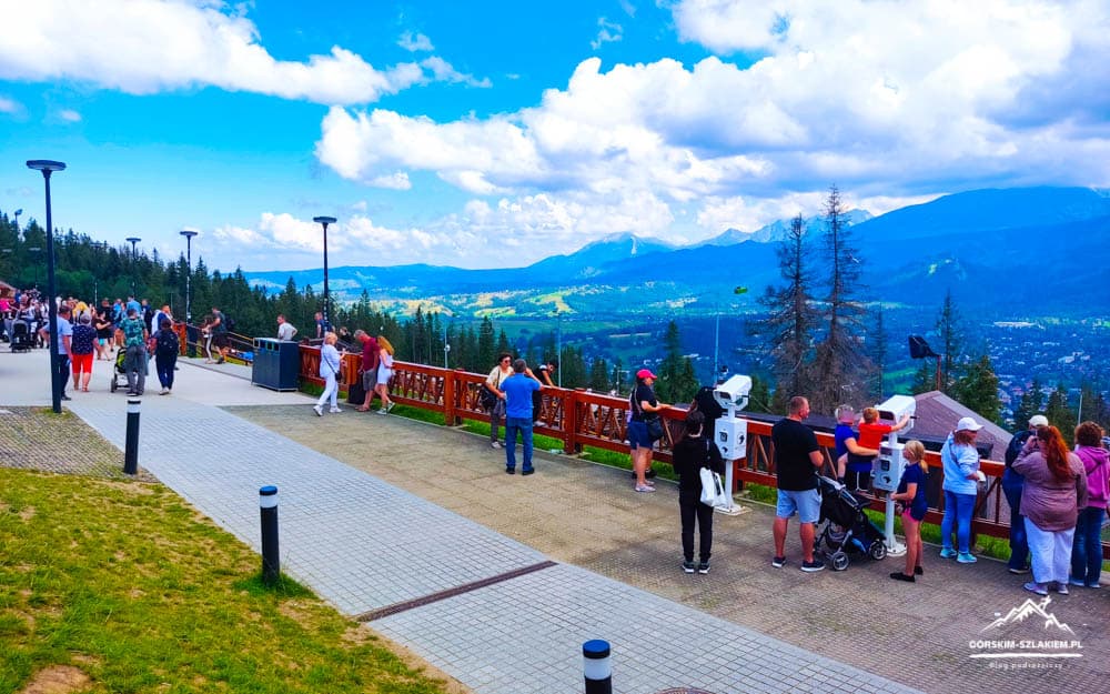 Crowd of visitors on a mountain viewpoint terrace overlooking a valley and forested hills under a blue sky.