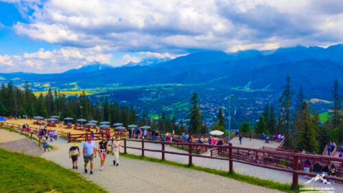 People walk along a paved path at a hilltop viewpoint with wooden rails and seating, overlooking a valley and distant mountains.