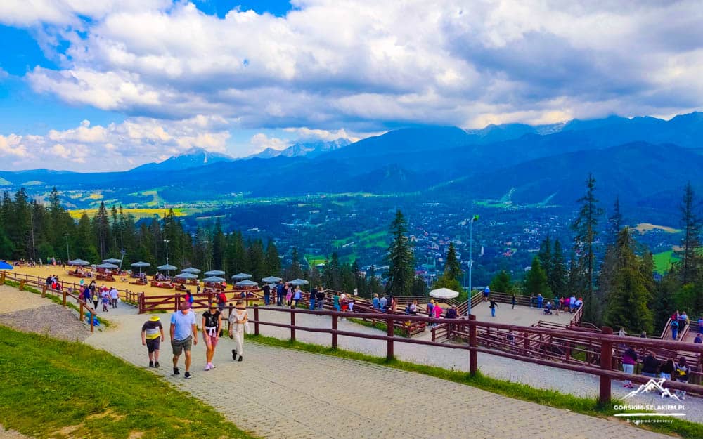 People walk along a paved path at a hilltop viewpoint with wooden rails and seating, overlooking a valley and distant mountains.