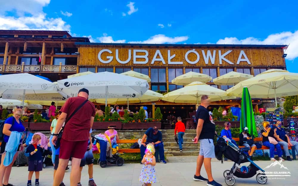 Outdoor cafe terrace in front of the Gubałówka building with white and yellow umbrellas and people enjoying the sunny day.