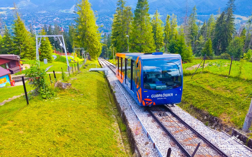Blue funicular car on a single-track railway winding through a green, forested hillside with distant buildings visible in the background.