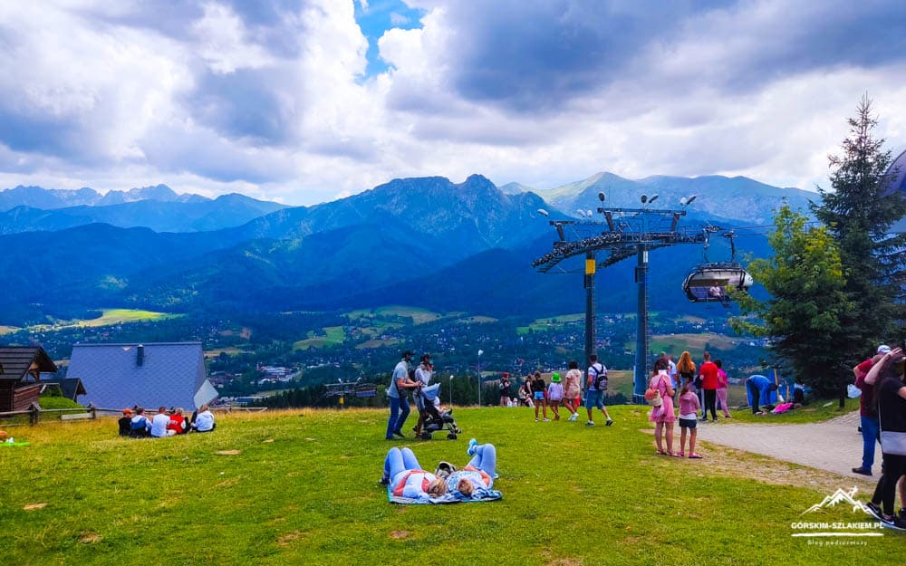 People relaxing on a grassy hillside with a chairlift and sweeping mountain valley beyond, under a cloudy blue sky.