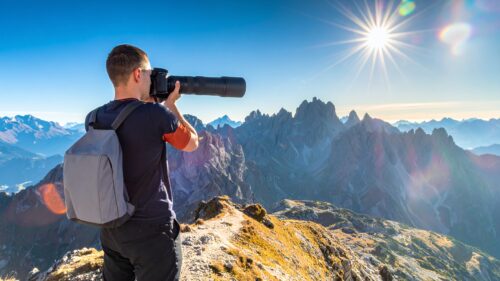 Photographer on a rocky mountain ridge aims a long telephoto lens toward distant peaks under a bright sun in a clear blue sky, backpack visible at back.