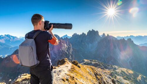 Photographer on a rocky mountain ridge aims a long telephoto lens toward distant peaks under a bright sun in a clear blue sky, backpack visible at back.