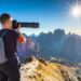 Photographer on a rocky mountain ridge aims a long telephoto lens toward distant peaks under a bright sun in a clear blue sky, backpack visible at back.